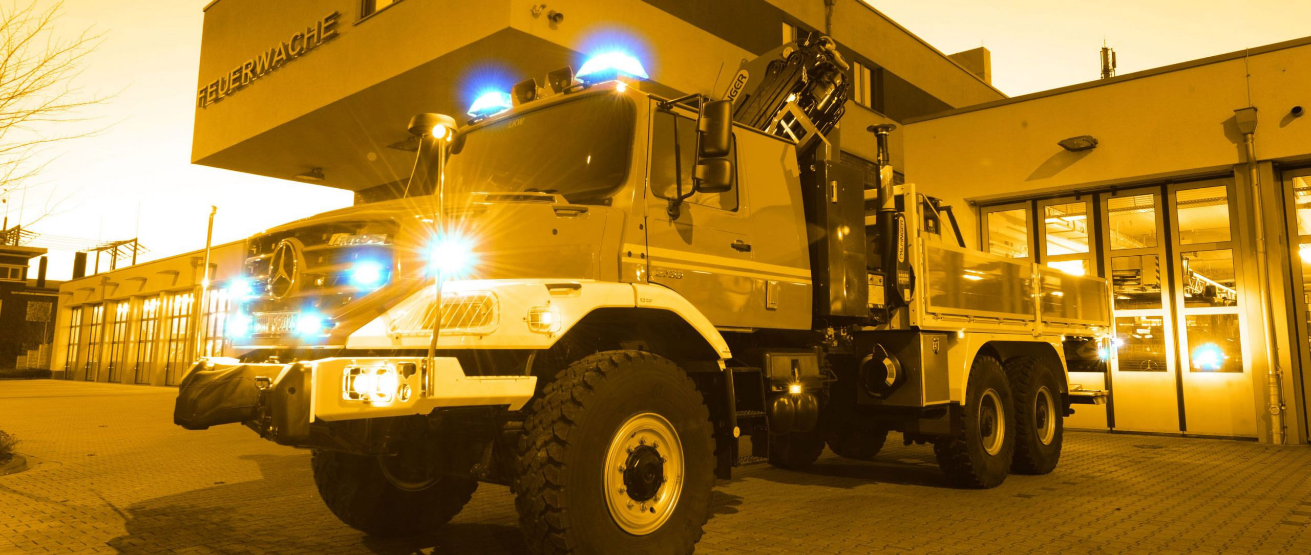 Vor einem Feuerwehrgebäude mit der Aufschrift „Feuerwache“ parkt ein Feuerwehrauto mit Blaulicht.