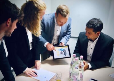 Vier Männer in Geschäftskleidung sitzen um einen Tisch herum, einer zeigt auf ein Tablet mit einer bunten Tabelle; auf dem Tisch stehen Wasserflaschen und Papiere.