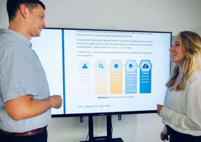 Zwei Personen stehen vor einem digitalen Bildschirm, auf dem ein Diagramm mit Text und Symbolen angezeigt wird, und sind in eine Diskussion oder Präsentation vertieft.