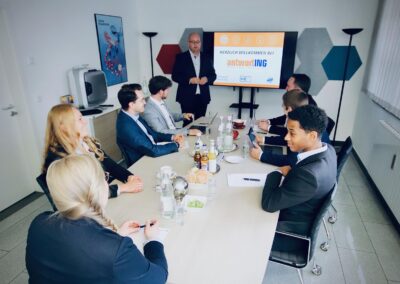 Eine Gruppe von Personen in Geschäftskleidung sitzt um einen Konferenztisch und hört einem Moderator zu, der neben einem Bildschirm steht, auf dem eine Begrüßungsnachricht für antwortING angezeigt wird.
