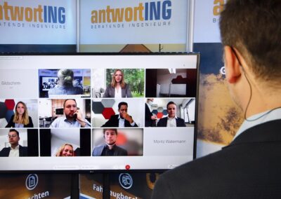 Ein Mann nimmt an einem virtuellen Meeting mit neun anderen Fachleuten teil, das auf einem großen Bildschirm in einer hell erleuchteten Büroumgebung sichtbar ist.