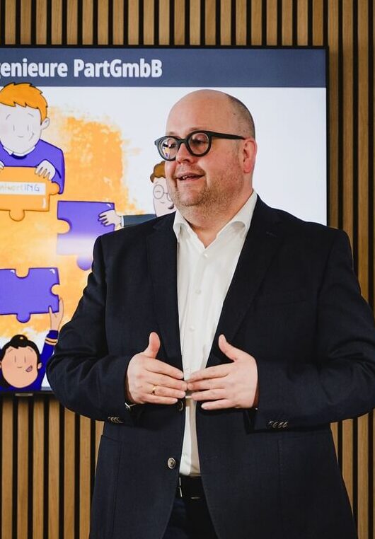 Ein Mann im Anzug h&auml;lt eine Pr&auml;sentation vor einem Bildschirm, auf dem eine R&auml;tselgrafik und deutscher Text angezeigt werden, w&auml;hrend im Vordergrund Menschen an einem Tisch sitzen.