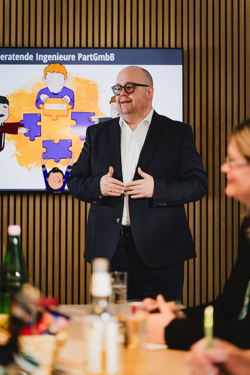 Ein Mann im Anzug hält eine Präsentation vor einem Bildschirm, auf dem eine Rätselgrafik und deutscher Text angezeigt werden, während im Vordergrund Menschen an einem Tisch sitzen.