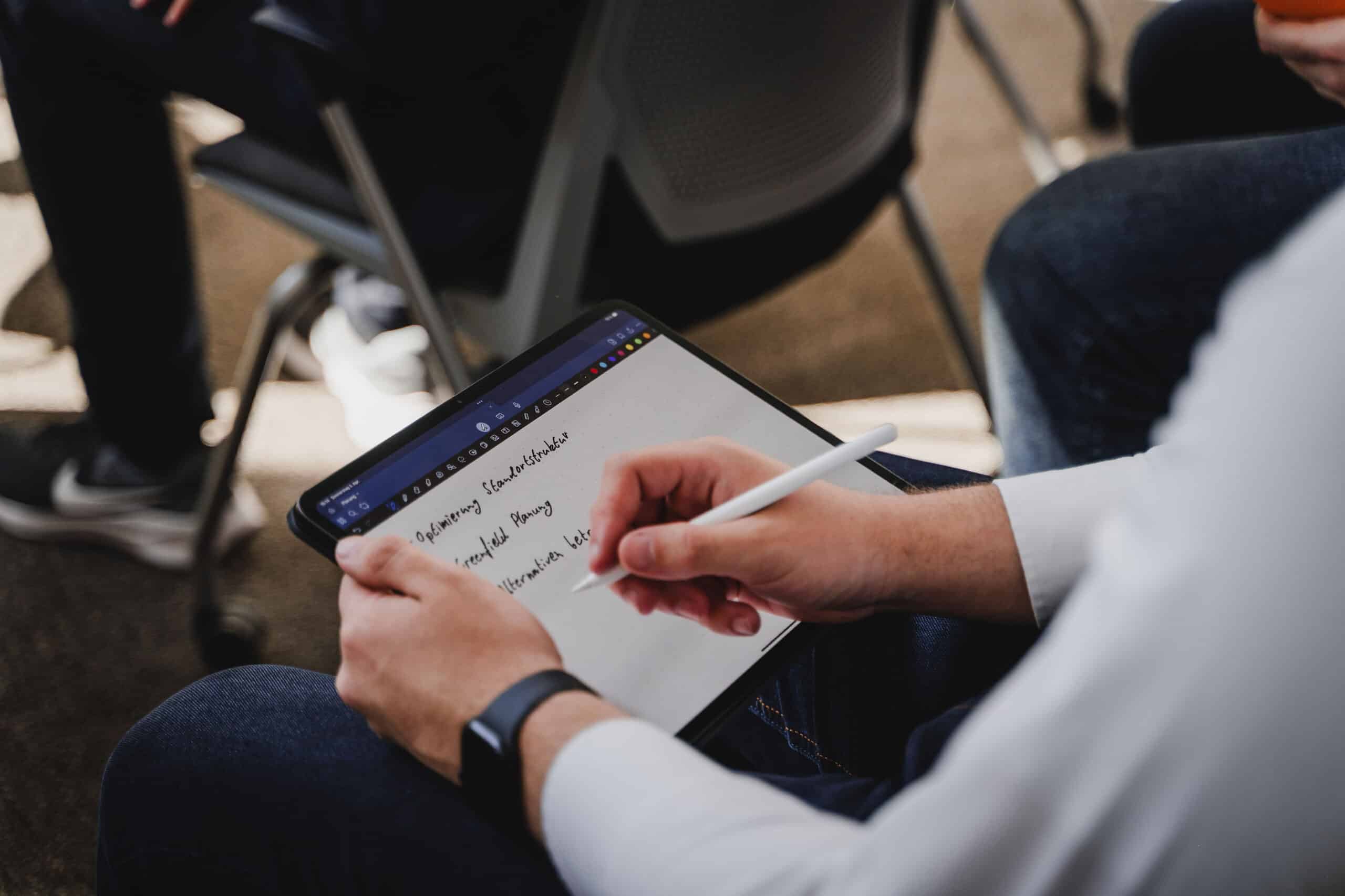 Eine Person macht im Sitzen mit einem Stift Notizen auf einem Tablet, im Hintergrund ist ein weiterer Stuhl zu sehen.