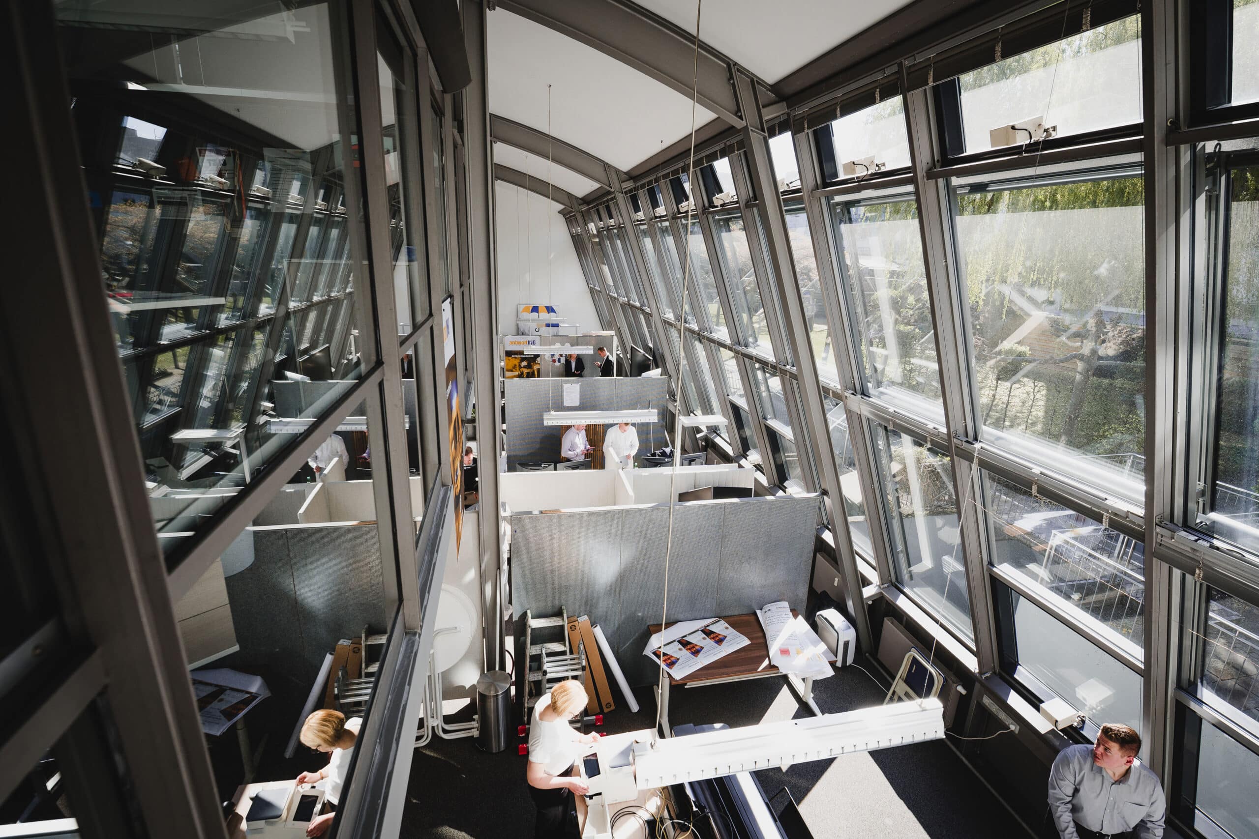 Großraumbüro mit großen Fenstern, mehreren Personen, die an Schreibtischen arbeiten, und hellem Tageslicht, das das moderne Interieur erhellt.