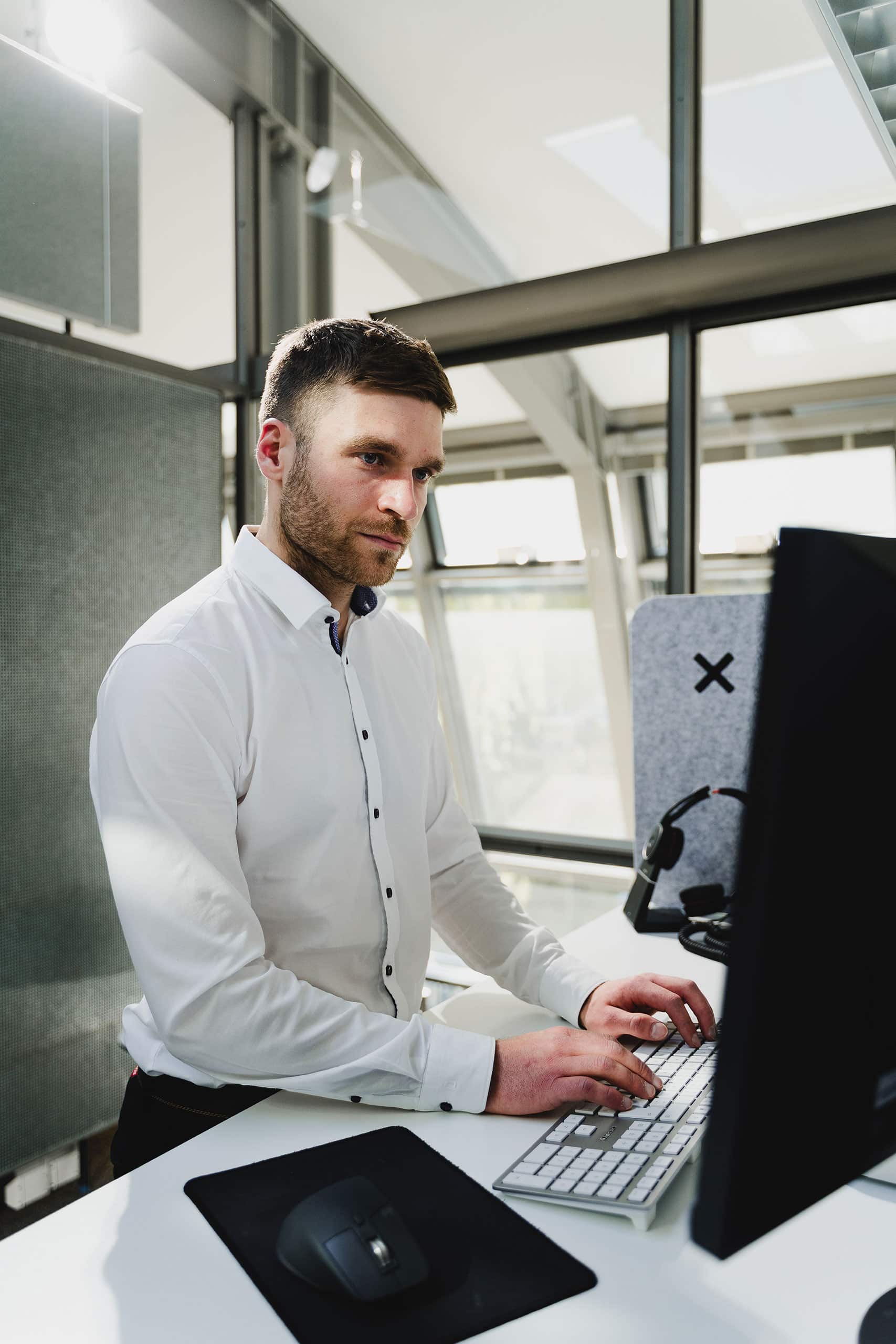 AntwortING_Stock-2404 Ein Mann im weißen Hemd steht an einem Schreibtisch, tippt auf einer Tastatur und schaut auf einen Computermonitor in einem modernen, gut beleuchteten Büro.