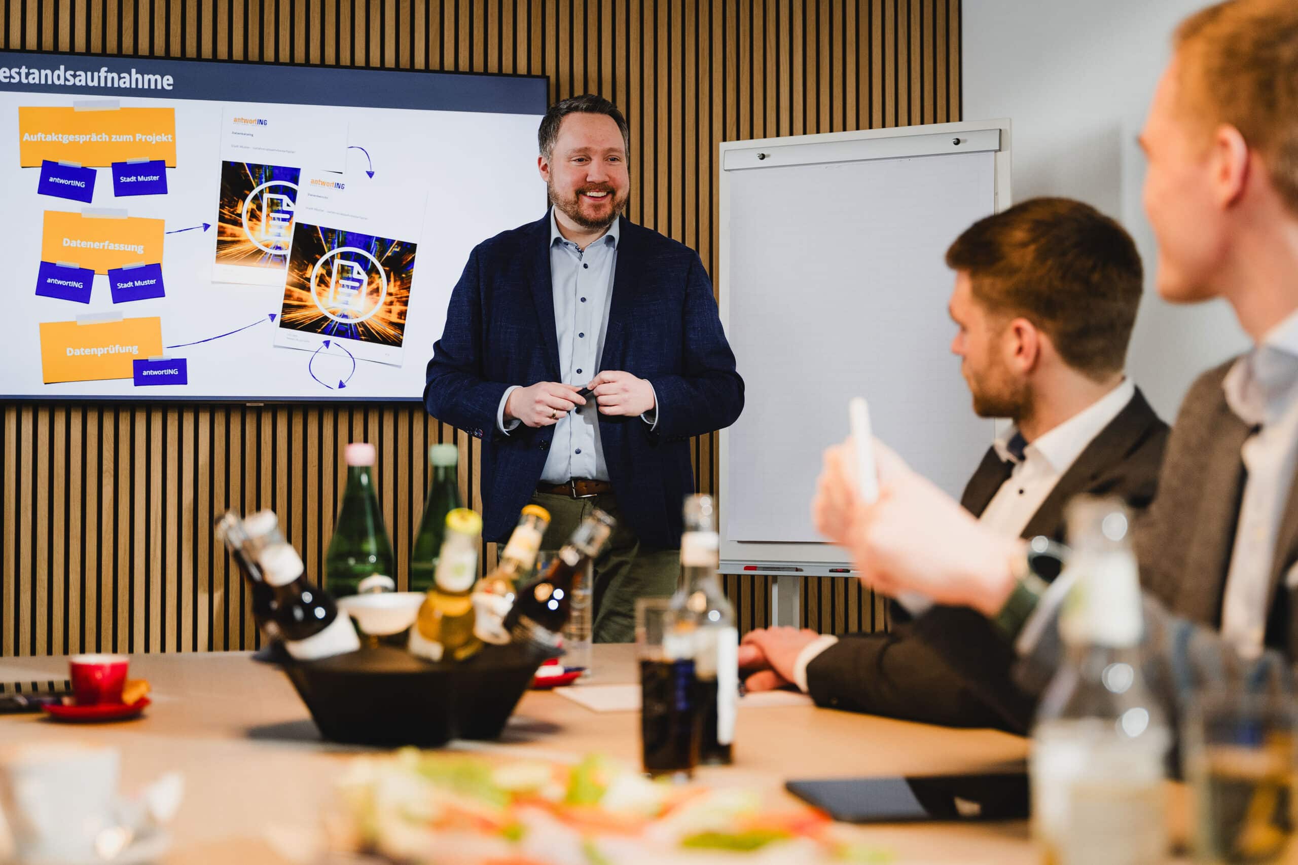 Ein Mann im Anzug hält vor drei sitzenden Kollegen in einem modernen Konferenzraum eine Präsentation. Auf dem Tisch stehen Getränke und Snacks.
