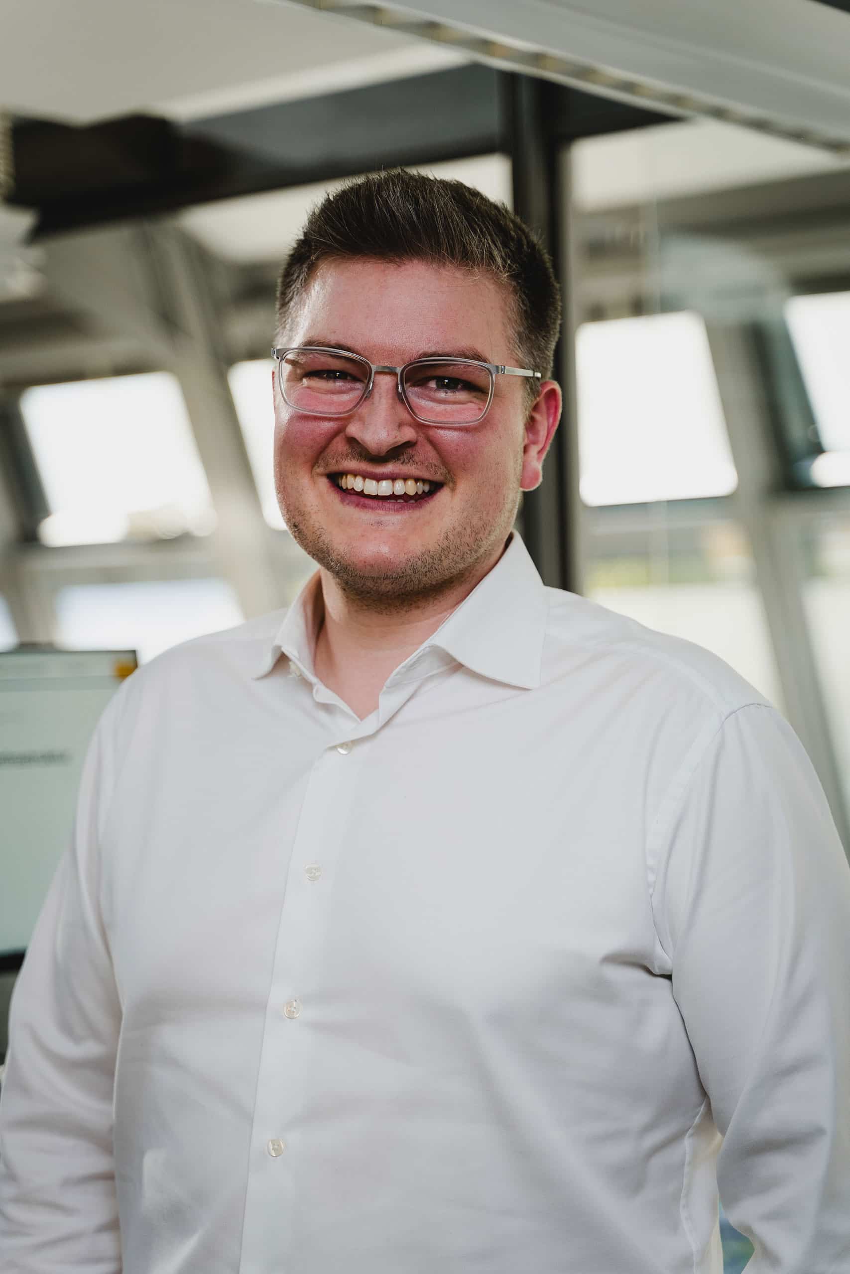 Ein Mann mit Brille und weißem Hemd steht lächelnd in einem gut beleuchteten, modernen Büroraum.