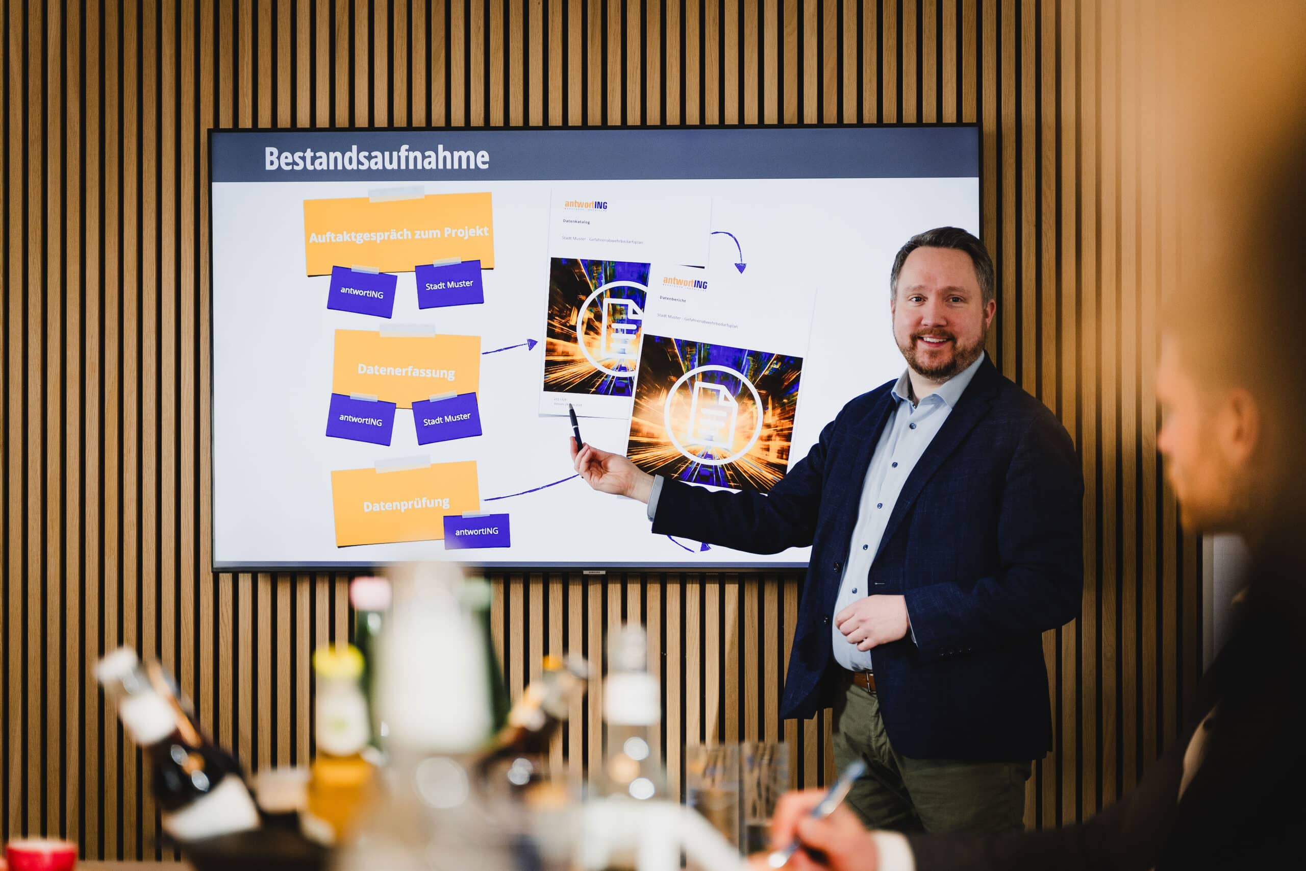 Ein Mann in Geschäftskleidung präsentiert in einem modernen Konferenzraum Folien mit Diagrammen und Schaubildern auf einer großen Leinwand.