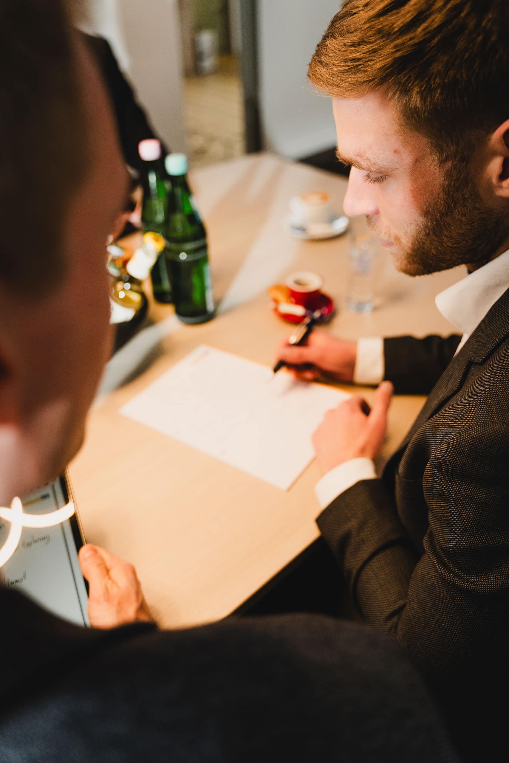 AntwortING_Stock-335 Zwei Männer in Anzügen sitzen mit einem Blatt Papier, Stiften, einer Flasche Wasser und Kaffeetassen an einem Schreibtisch und scheinen gemeinsam Dokumente zu besprechen oder durchzugehen.