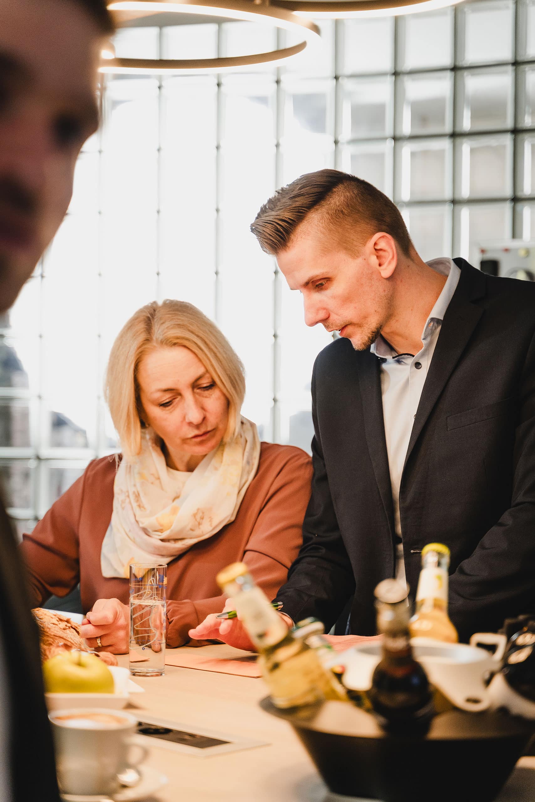 Zwei Personen in Businesskleidung schauen gemeinsam auf ein Smartphone an einem Tisch, im Vordergrund stehen Getränke, Essen und Flaschen.