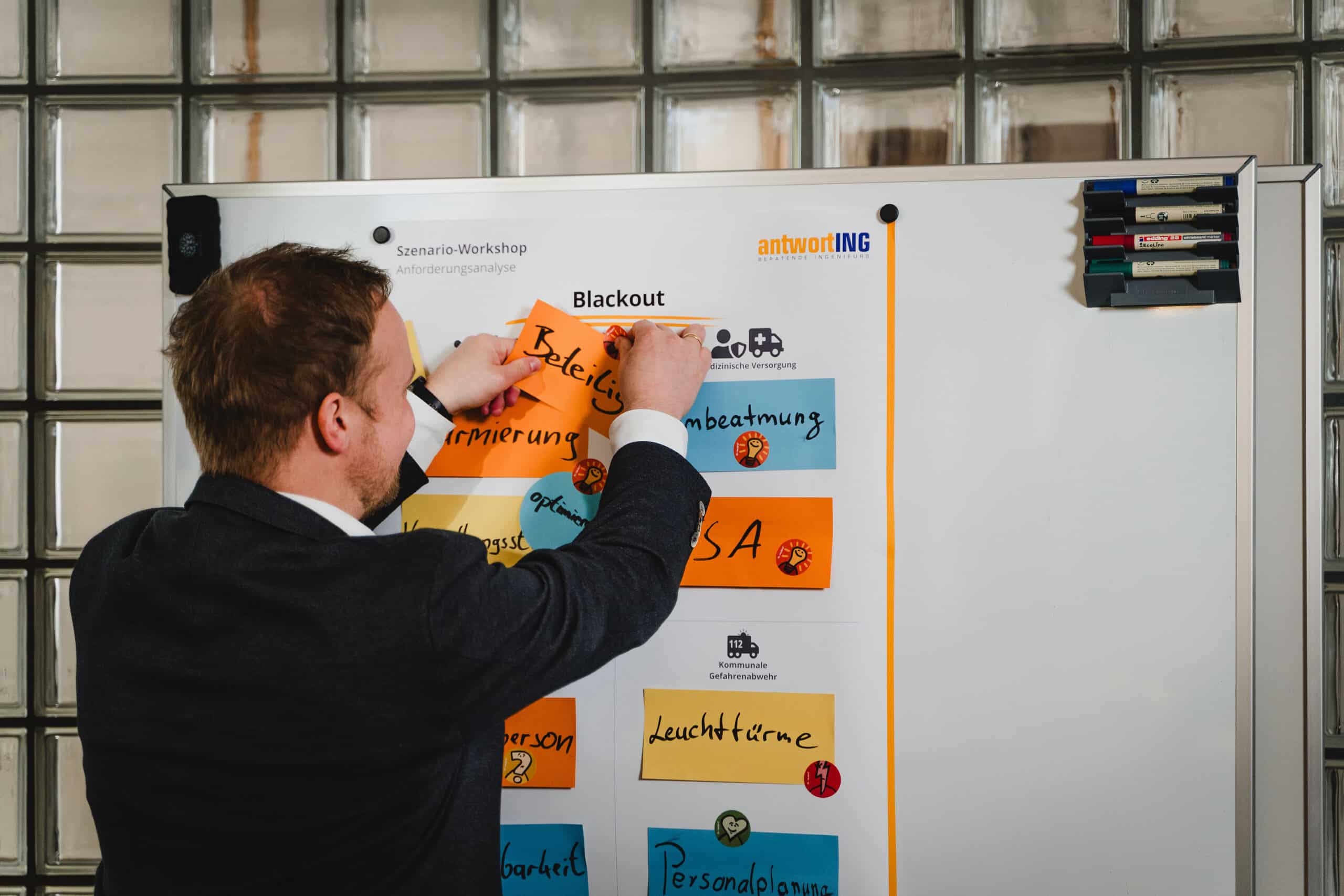 In einer modernen Büroumgebung befestigt ein Mann einen Haftzettel an einem Whiteboard, das mit bunten Notizen und Workshop-Themen, darunter „Blackout“, bedeckt ist.