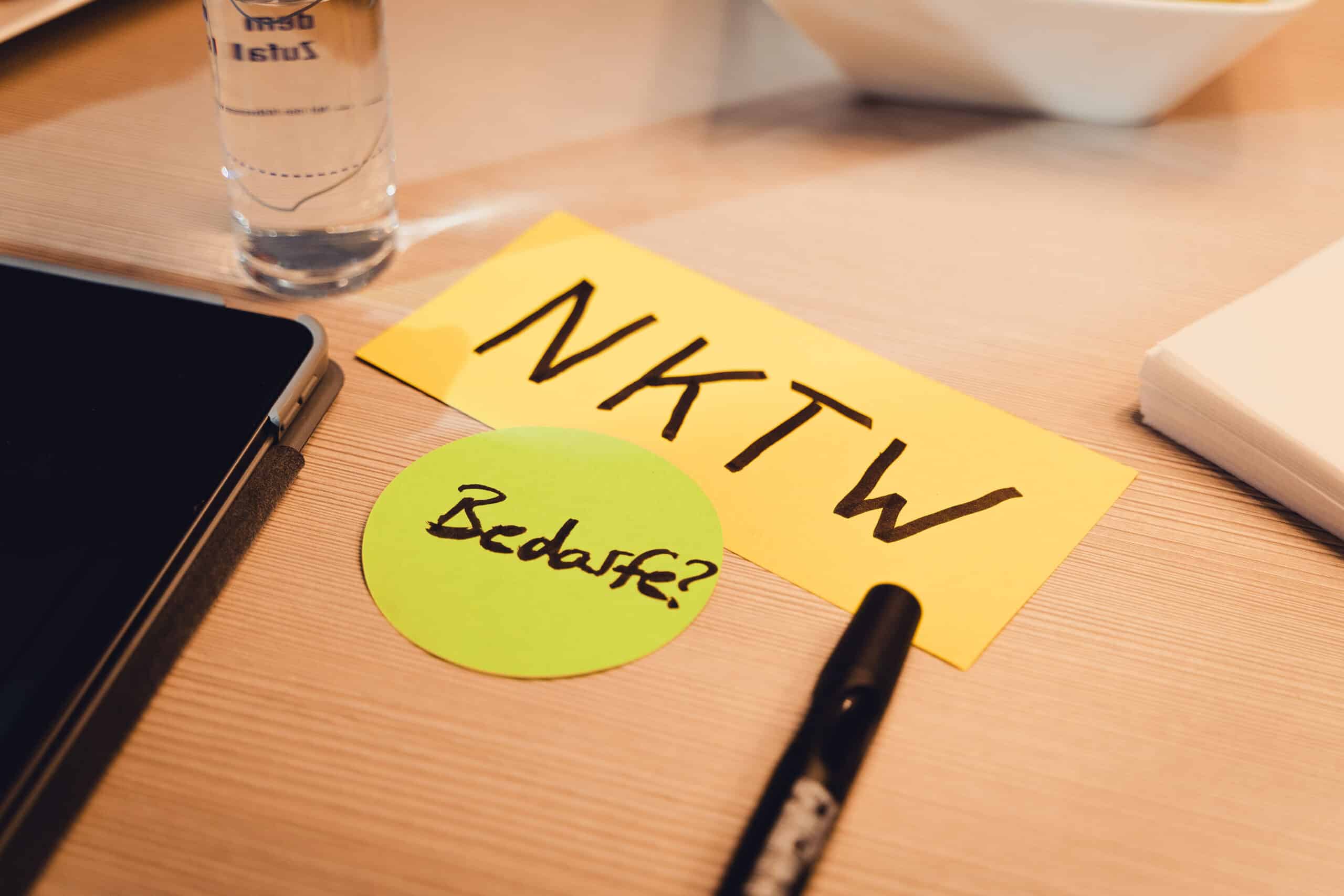 Eine gelbe Karte mit der Aufschrift „NKTW“ und ein grüner Kreis mit der Aufschrift „Bedarf?“ liegen auf einem Tisch neben einem schwarzen Stift, einem Tablet und einem Glas Wasser.