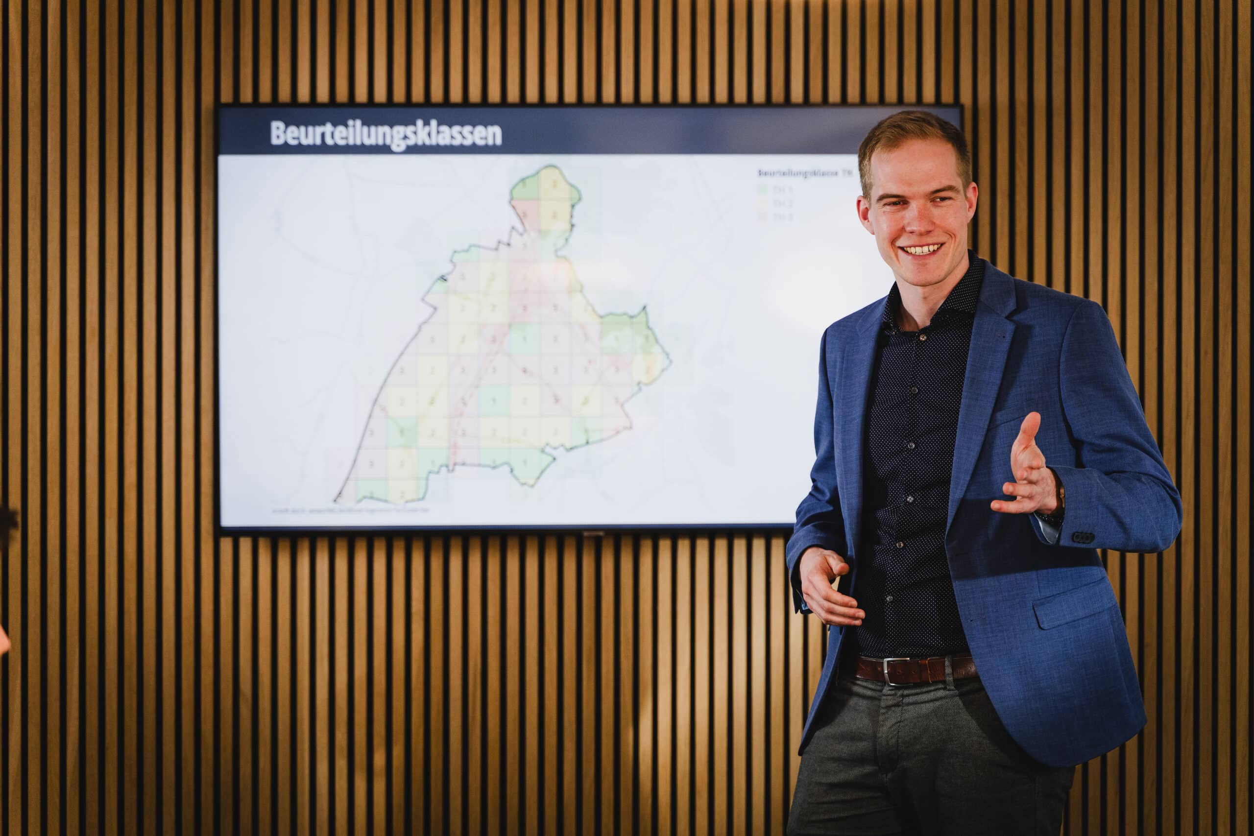 Ein Mann in einem blauen Blazer präsentiert vor einem Bildschirm eine Karte mit der Aufschrift „Beurteilungsklassen“ auf Deutsch.