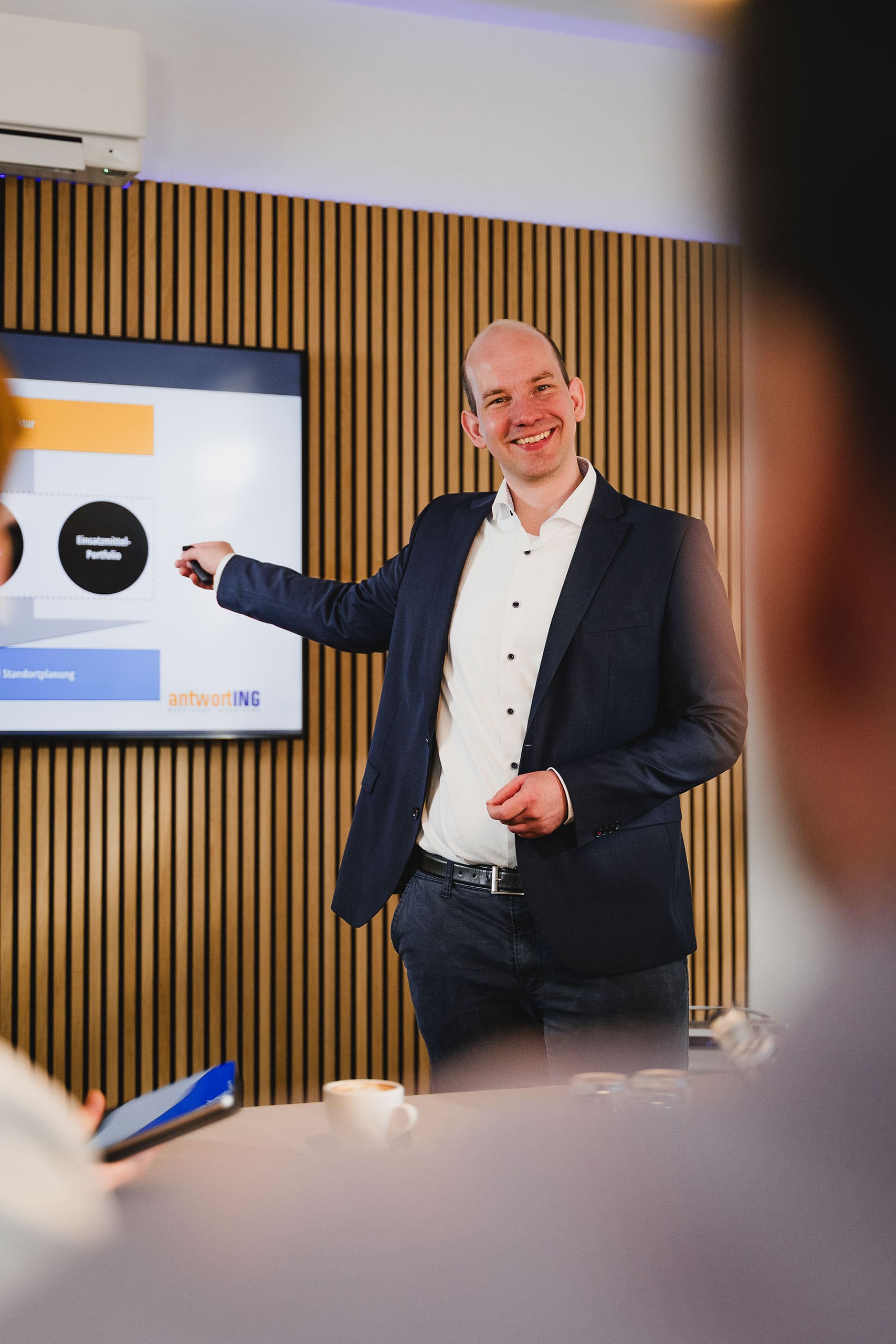 AntwortING_Stock-767 Ein Mann im Anzug steht vor einem Bildschirm und hält eine Präsentation, wobei er auf ein Diagramm mit Text und Grafiken zeigt.