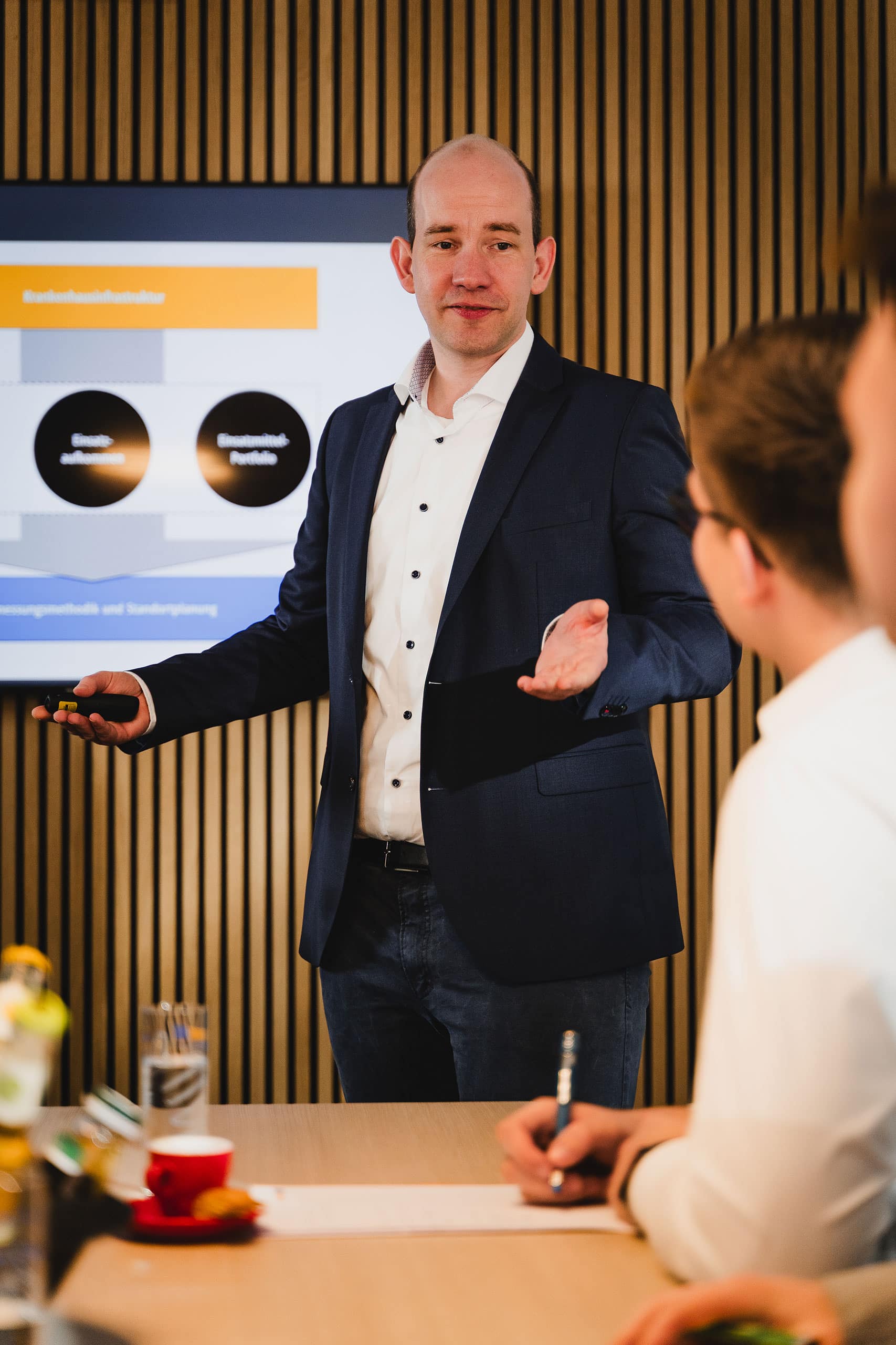Ein Mann im Anzug hält vor zwei sitzenden Personen eine Präsentation und gestikuliert dabei mit einer Hand. Im Hintergrund werden auf einem Bildschirm Diagramme und Text angezeigt.