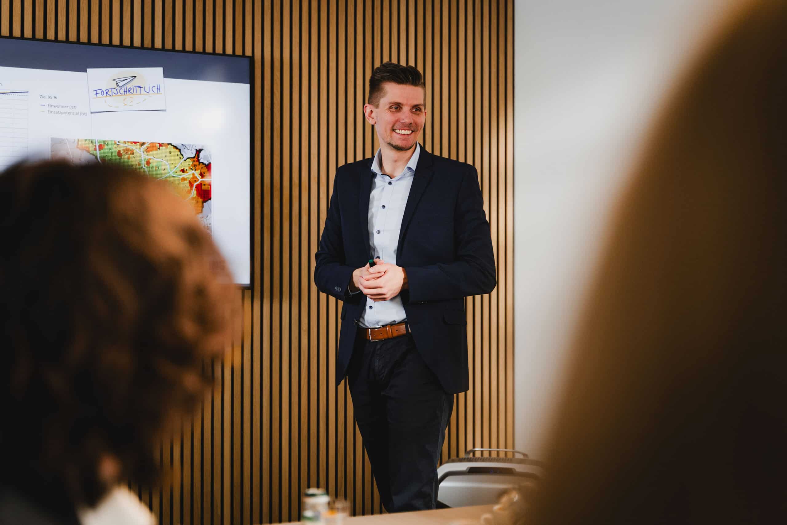 Ein Mann im Anzug steht l&auml;chelnd vor einem Pr&auml;sentationsbildschirm, w&auml;hrend er in einer modernen B&uuml;roumgebung eine Rede vor einer Gruppe h&auml;lt.