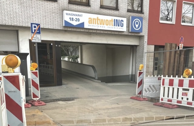 Einfahrt zu einem Bürogebäude am Waidmarkt 18–20 mit einem großen Firmenschild von „antwortING – Beratende Ingenieure“ über der Garageneinfahrt. Auf dem Bürgersteig davor befinden sich Absperrungen und Baustellenleuchten. Rechts und links sind Teile angrenzender Gebäude zu sehen. Das Gebäude besteht aus heller Klinkerfassade mit mehreren Fenstern.