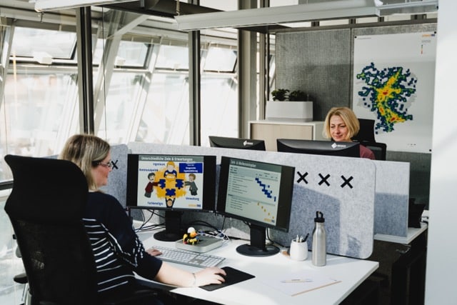 Zwei Frauen arbeiten an Computern in einem modernen, lichtdurchfluteten Großraumbüro. Im Vordergrund sitzt eine Frau mit Brille an einem Arbeitsplatz mit zwei Monitoren, auf denen Präsentationsgrafiken zu Digitalisierung und Technologie angezeigt werden. Im Hintergrund arbeitet eine weitere Frau an einem separaten Schreibtisch. Zwischen den Plätzen befinden sich graue Akustiktrennwände mit schwarzen „X“-Markierungen. Eine farbige Deutschlandkarte hängt an der Wand, daneben stehen Pflanzen auf einem Schrank.
